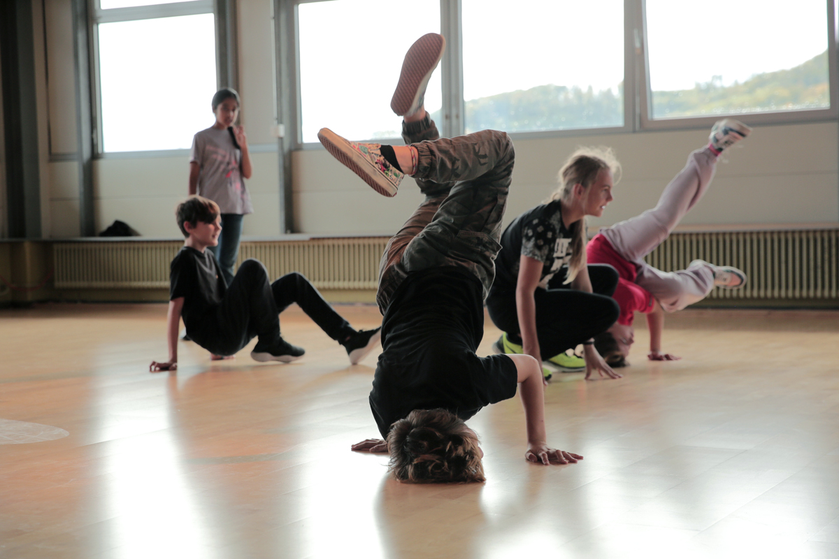 Kinder machen Breakdance Bewegungen und Posen, wie einen seitlichen Kopfstand, in einer Turnhalle