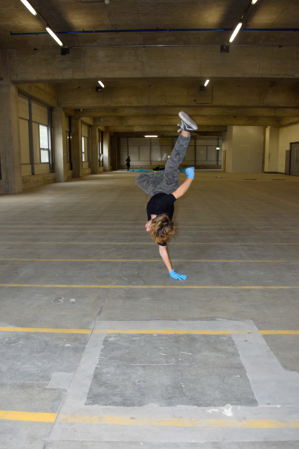Ein Kind macht in einer Parkhaus einen einarmigen Handstand und trägt hellblaue Handschuhe