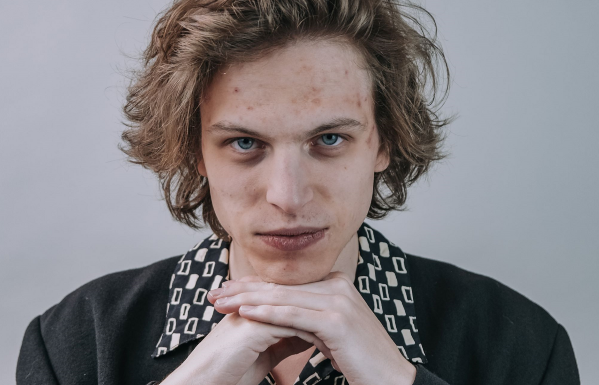 Portrait von Vili Polajnar, der vor grauem Hintergrund in schwarzem Jackett und gemustertem Hemd sein Kinn auf seinen Händen abstützt und frech in die Kamera schaut