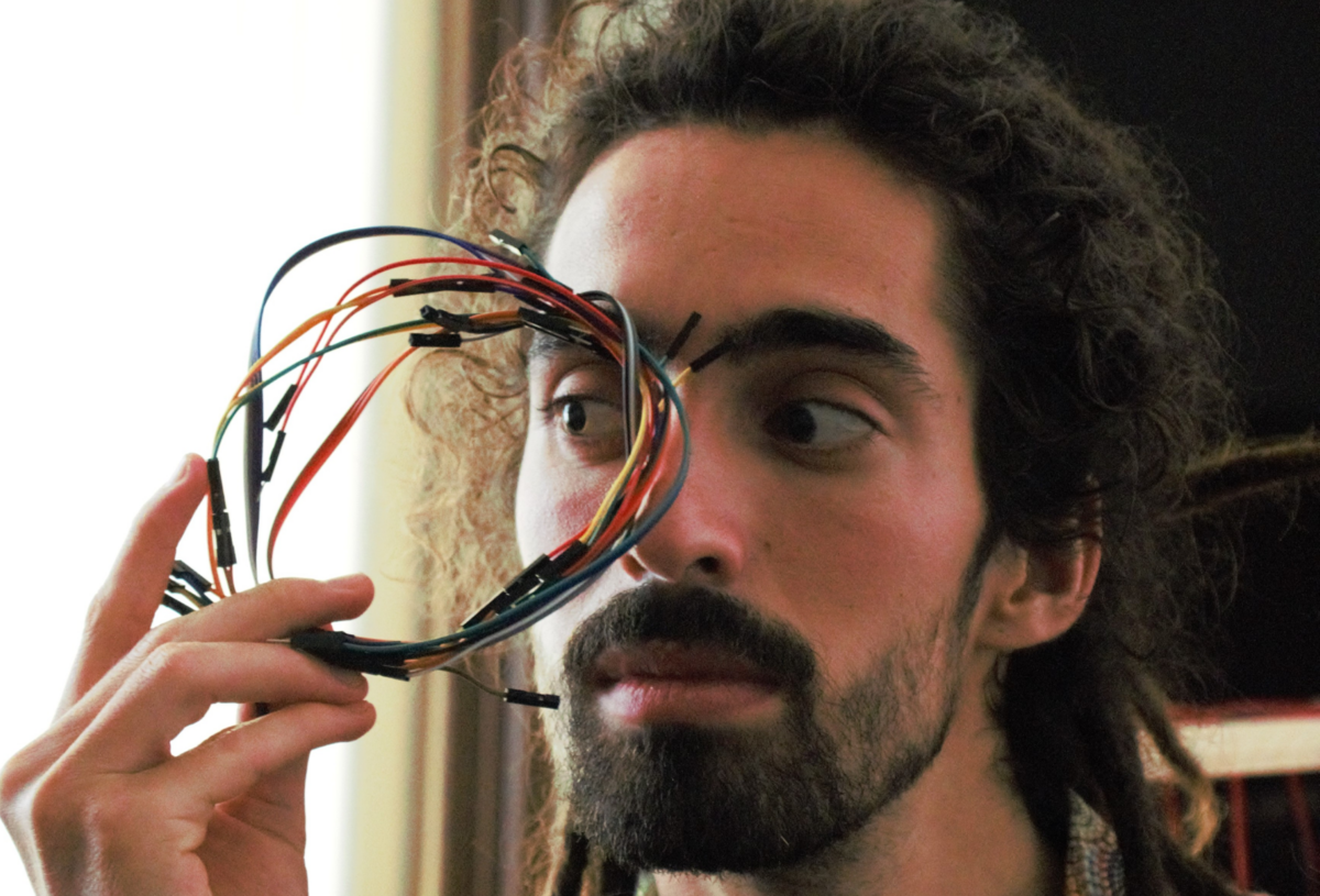 Alexandre Tavares hält sich einen Kreis aus Elektrokabeln vor sein eines Auge, durch das er schräg hindurch schaut.