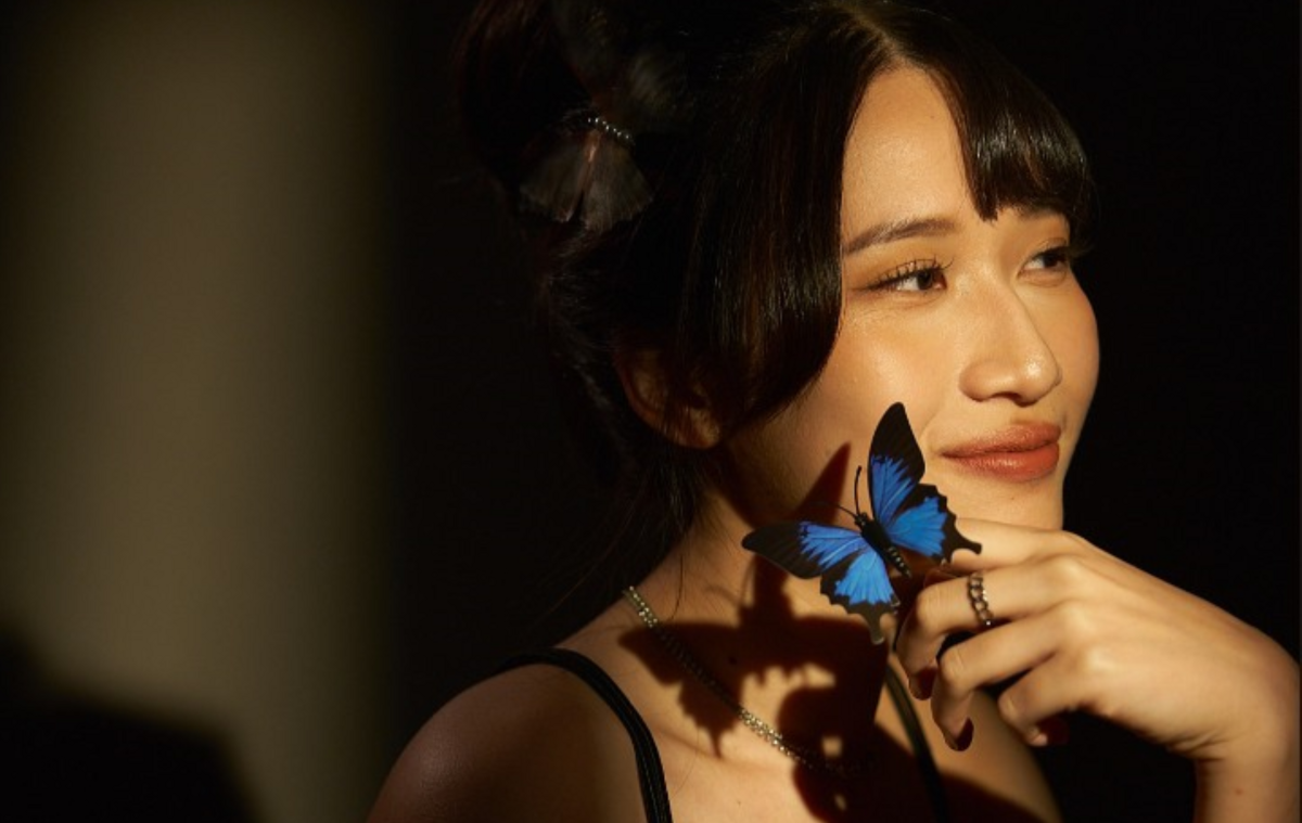 Yu-Yun Peng befindet sich in einem sehr dunklen Raum, während ein Lichtstrahl genau auf sie trifft, sie stützt ihr Kinn in einer Hand, auf dem ein blauer Schmetterling sitzt und schaut aus dem Bild raus.