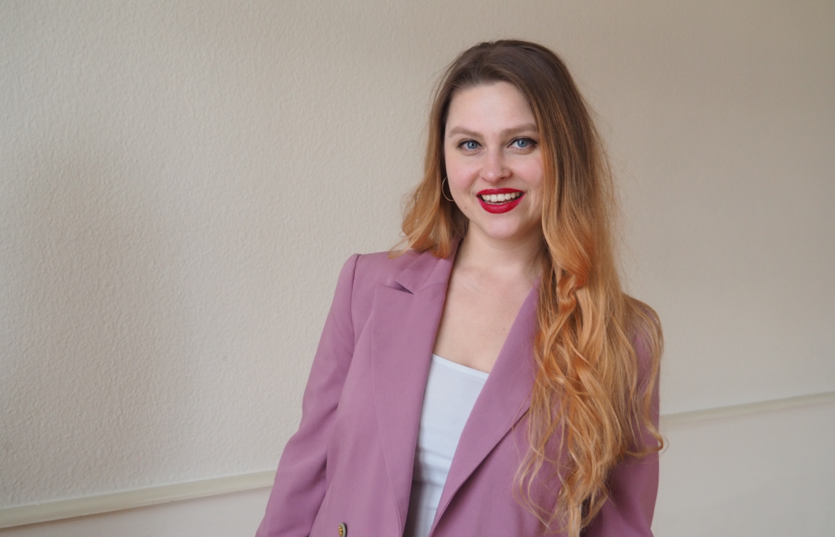 Barbora Jirásková steht vor einem weiss neutralen Hintergrund, von dem sich ihr rosa farbenes Jackett abhebt und lacht sympathisch in die Kamera