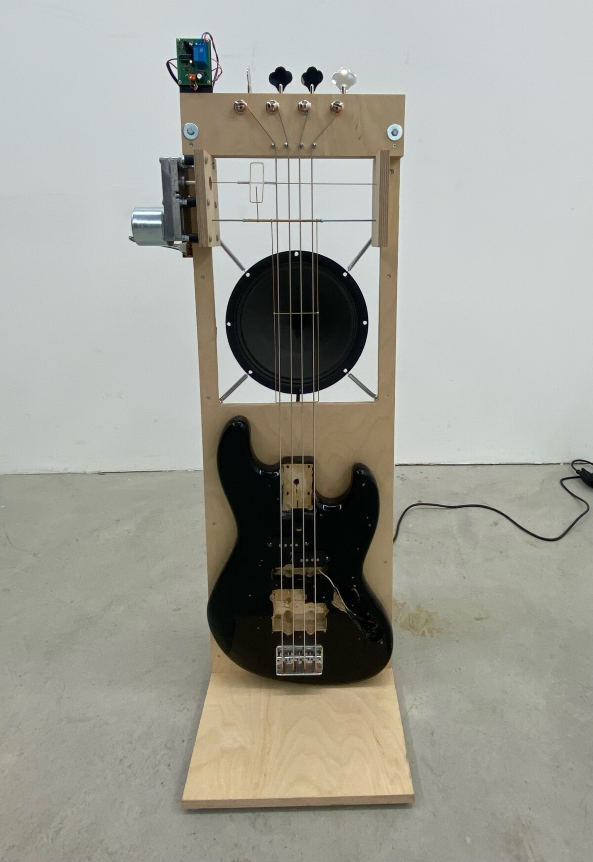 Die Installation Bass-Automated, der Holzkörper eines elektrischen Basses ist an einer senkrecht stehenden Holzplatte befestigt, die Saiten des Bass sind oben technisch präpariert, sodass sie durch Technik und deshalb automatisch gespielt werden.