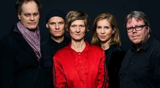 Foto des Eva Kruse Quintet, die in schwarzer Kleidung vor einem schwarzen Hintergeund rund um Eva Kruse, die in rot gekleidet ist, stehen