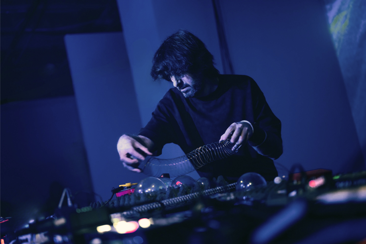 Foto von Fernando Manassero, der in blauem Bühnenlicht eine auseinanderziehbare Metallspirale über sein elektronisches Musikset hält
