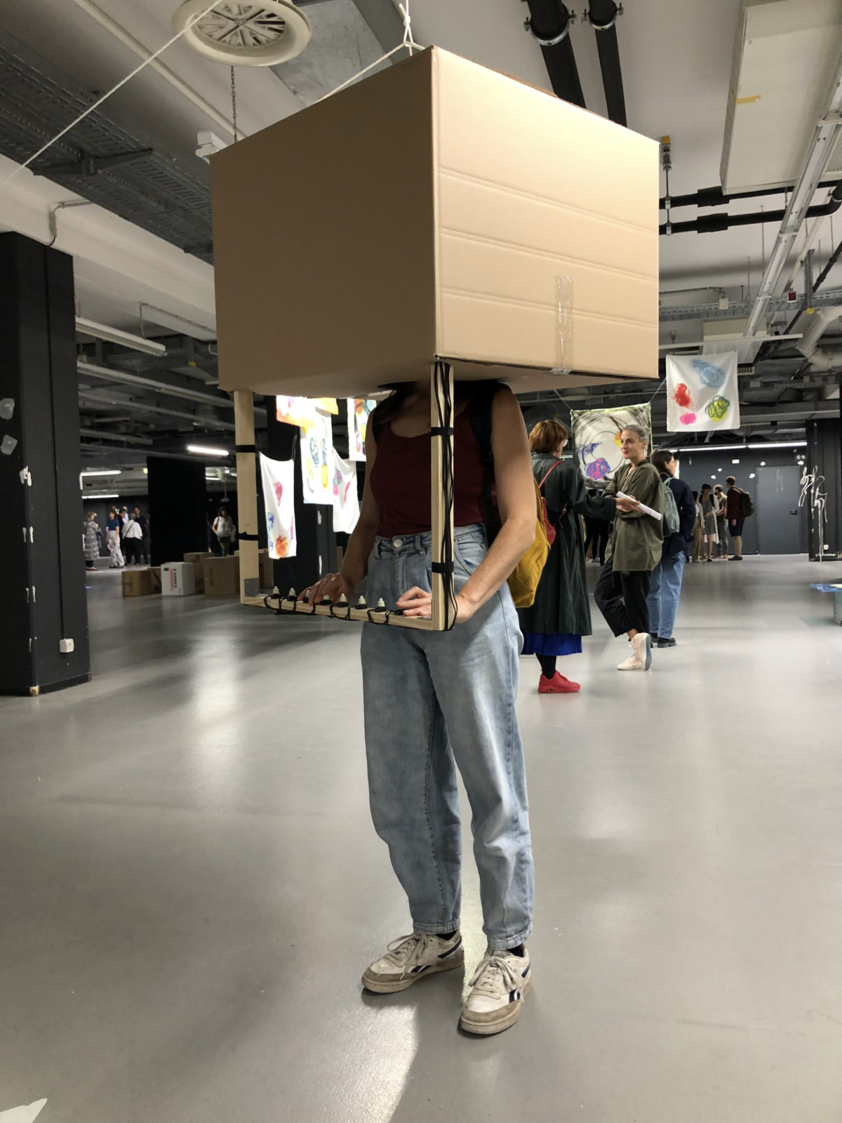 Eine person trägt hält ihren Kopf in eine grosse Kartonschachtel, die von der Decke hängt und bedient Knöpfe, die sich auf Hüft Höhe befinden und mit dem Karton verbunden sind.