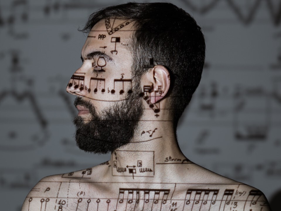 Das Foto zeigt Miguel Ángel García Martín, der mit geschlossenen Augen und nacktem Oberkörper vor einer Leinwand steht, auf die Musiknoten projiziert werden, die deshalb auch auf ihn projiziert werden.