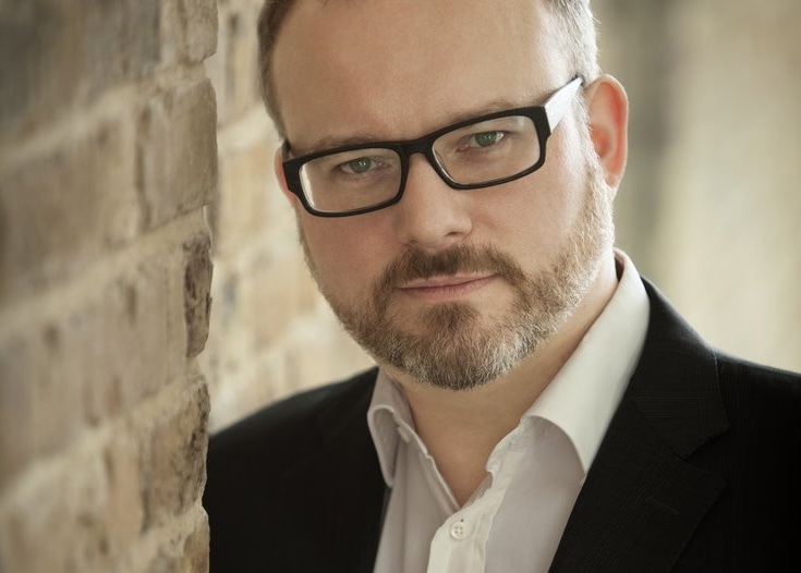 Portrait von Nicolas Hodges, der hinter einer Steinwand hervorschaut
