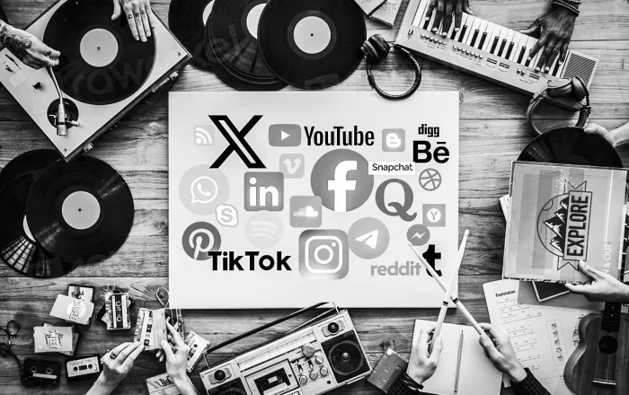 schwarz weisses Bild, mit einer Kollektion an verschiedenen Musik Medien, wie einem Plattenspieler, ein Radio, ein Keyboard oder Kassetten, die rund um einen Laptop mit den Logos vieler sozialer Medien platziert sind